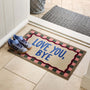 A rectangular doormat with the words “LOVE YOU, BYE” in blue sits on a tiled entryway floor next to a pair of blue sneakers. The mat has a beige center, a pink and green checkered border, and a dark red wavy inner border.