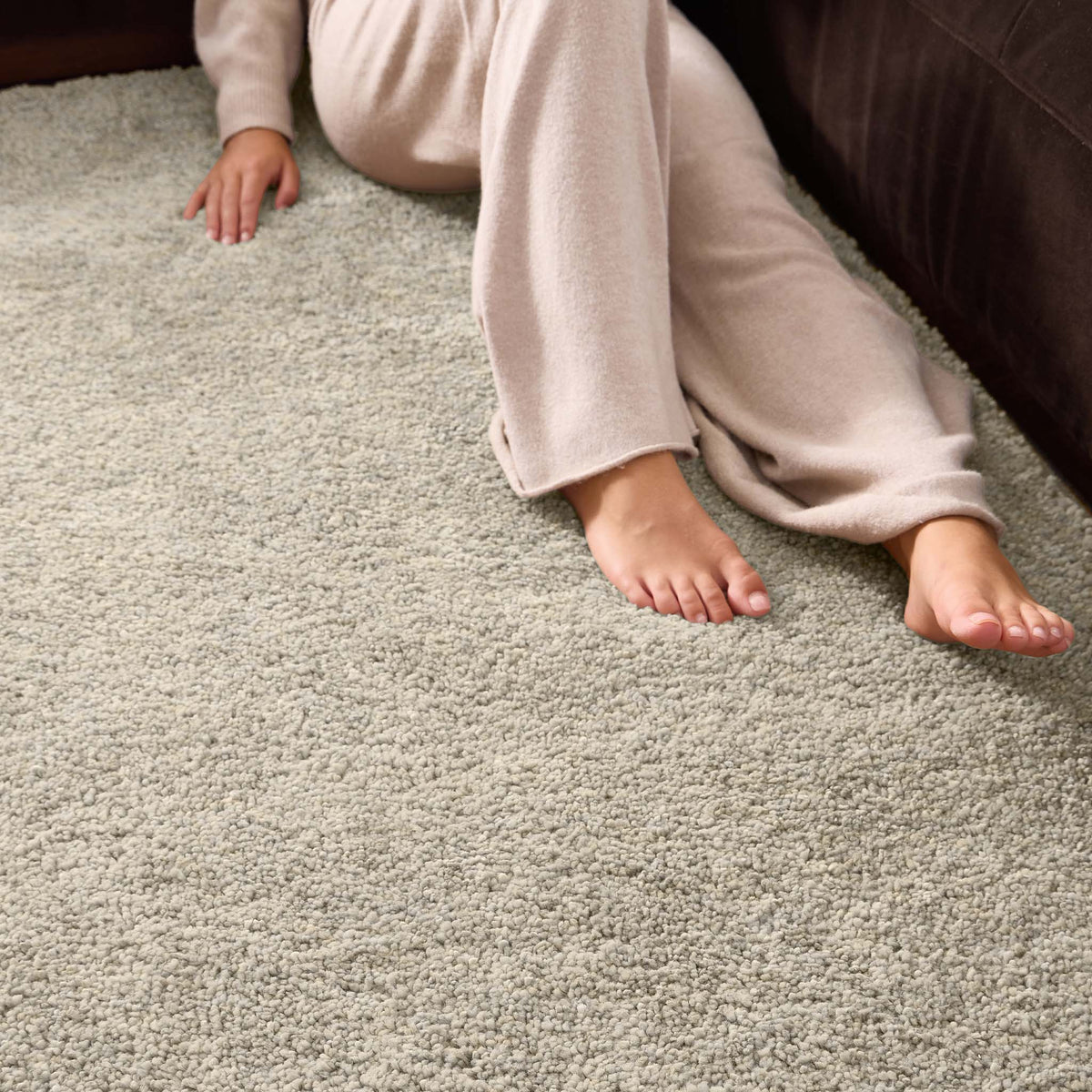 A close-up of a person sitting on a light beige shag rug next to a dark brown sofa, wearing beige pajama pants and bare feet.