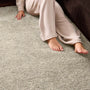 A close-up of a person sitting on a light beige shag rug next to a dark brown sofa, wearing beige pajama pants and bare feet.