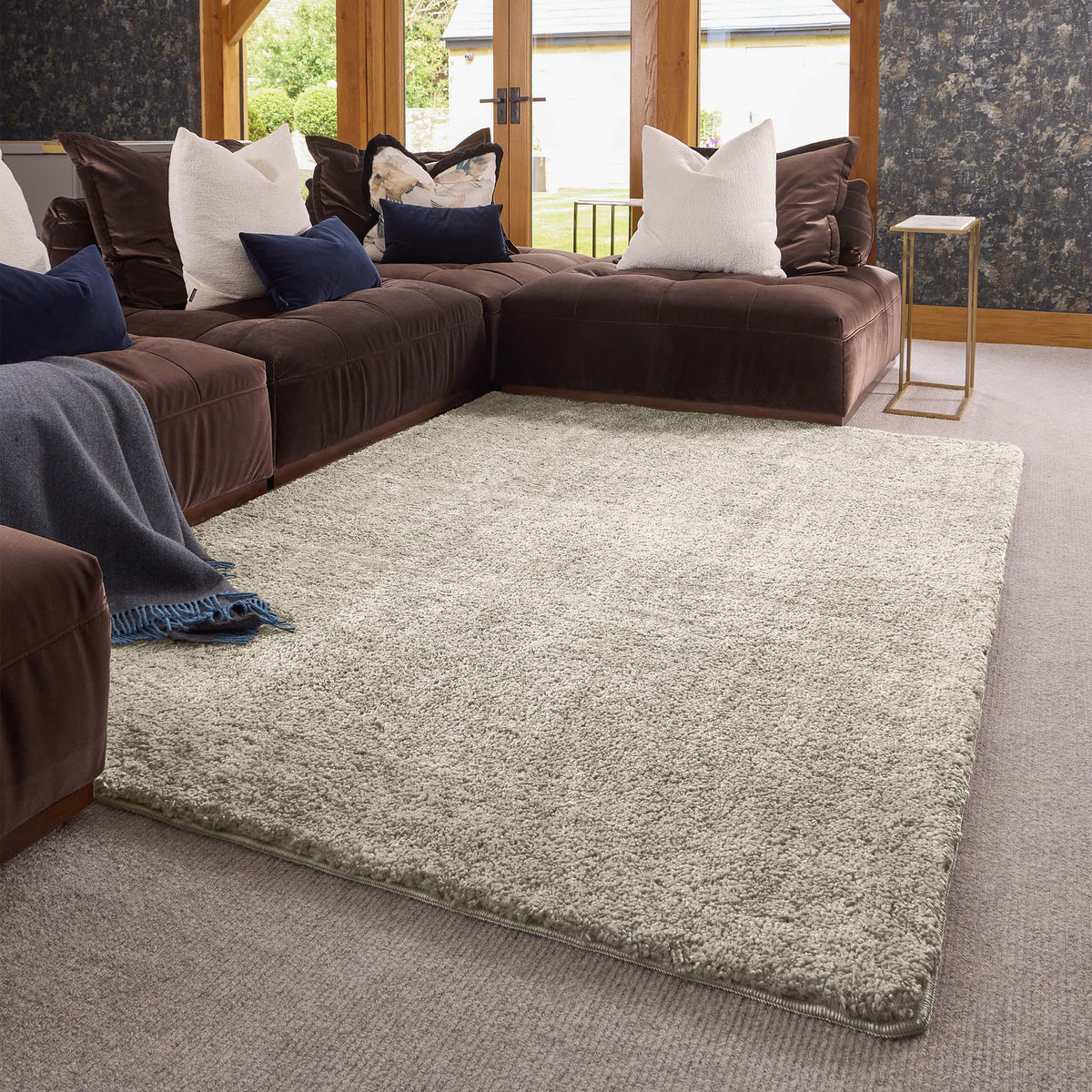 A large rectangular shag area rug in a light beige-gray color, styled in a modern living room setting with a dark brown sectional sofa, decorative pillows, and a blue throw blanket.