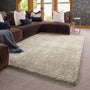 A large rectangular shag area rug in a light beige-gray color, styled in a modern living room setting with a dark brown sectional sofa, decorative pillows, and a blue throw blanket.