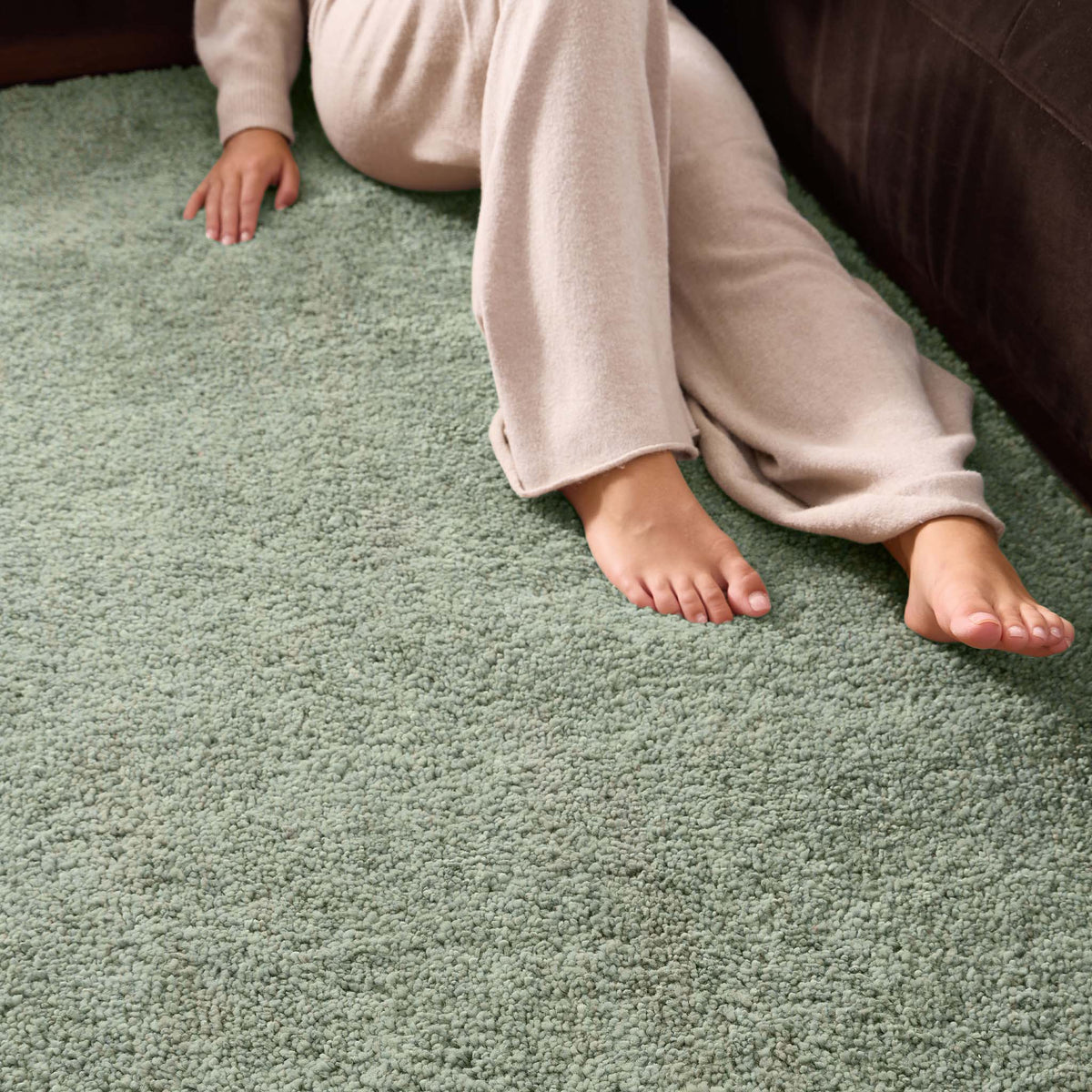 A person wearing beige lounge pants sits on a textured sage green shag rug next to a dark brown sofa, with their bare feet visible.