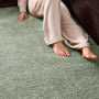 A person wearing beige lounge pants sits on a textured sage green shag rug next to a dark brown sofa, with their bare feet visible.