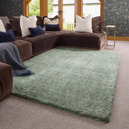 A large rectangular shag area rug in a muted sage green color, styled in a modern living room setting with a dark brown sectional sofa, decorative pillows, and a light-colored carpet.