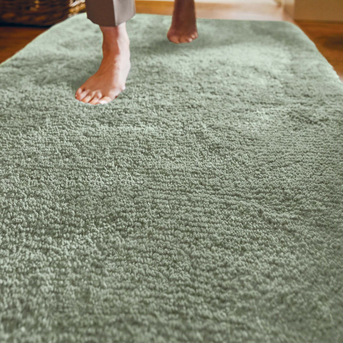 A close-up of a pale sage green shag rug with bare feet stepping onto it in a home setting.