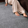 A person wearing beige lounge pants sits on a textured gray shag carpet next to a dark brown sofa, with their bare feet and hand visible.