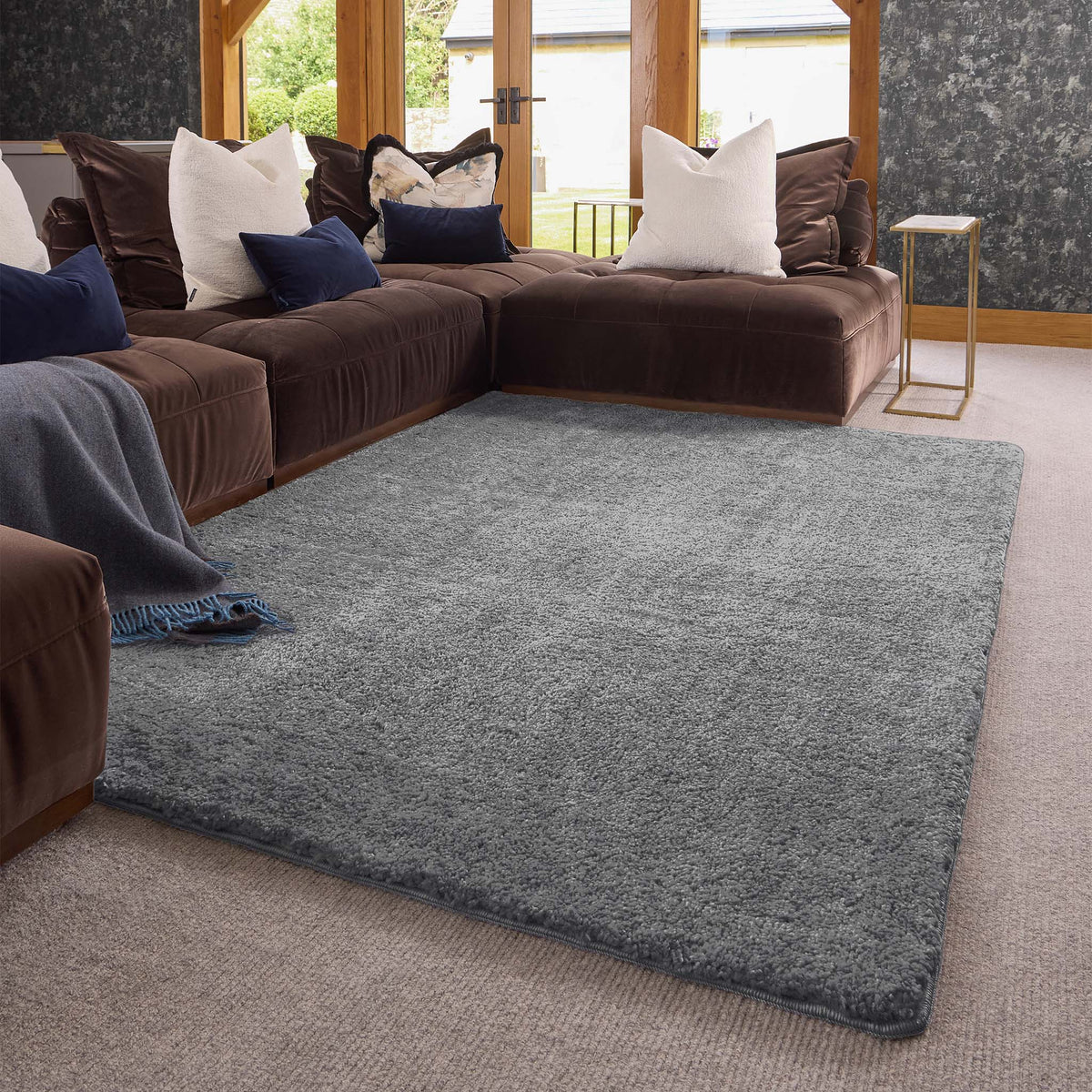A large rectangular gray shag area rug styled in a modern living room setting with a brown sectional sofa, white and navy pillows, and a small gold side table.