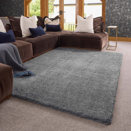 A large rectangular gray shag area rug styled in a modern living room setting with a brown sectional sofa, white and navy pillows, and a small gold side table.