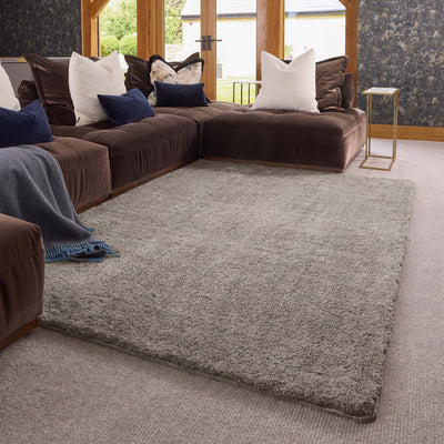 A large, light stone shag area rug styled in a modern living room setting with a brown sectional sofa, decorative pillows, and a small side table.