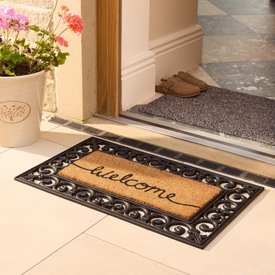 A rectangular welcome doormat with a black ornate scroll border and natural tan coir center featuring the word 'welcome' in black script, placed in front of a doorway beside a potted flower.