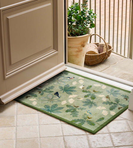 A rectangular entryway doormat with a green border and a leafy botanical pattern in muted green, beige, and blue tones, placed in front of an open door on a tiled floor with a potted plant and basket visible outside.