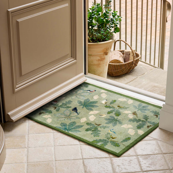 A rectangular entryway doormat with a green border and a leafy botanical pattern in muted green, beige, and blue tones, placed in front of an open door on a tiled floor with a potted plant and basket visible outside.