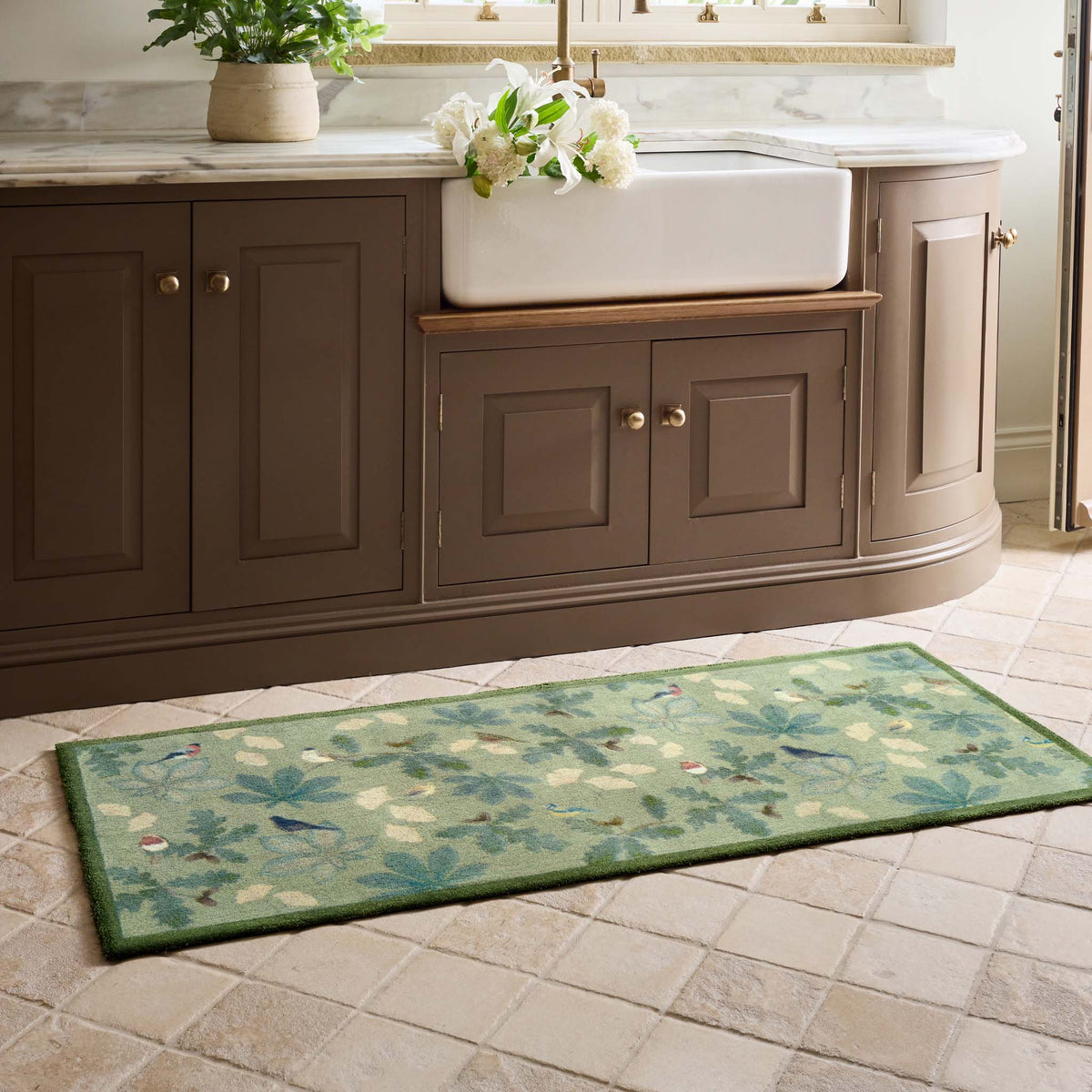 A rectangular green runner with a botanical bird pattern lies on a beige tiled floor in front of a brown vanity cabinet with a white farmhouse sink.