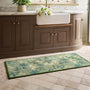 A rectangular green runner with a botanical bird pattern lies on a beige tiled floor in front of a brown vanity cabinet with a white farmhouse sink.