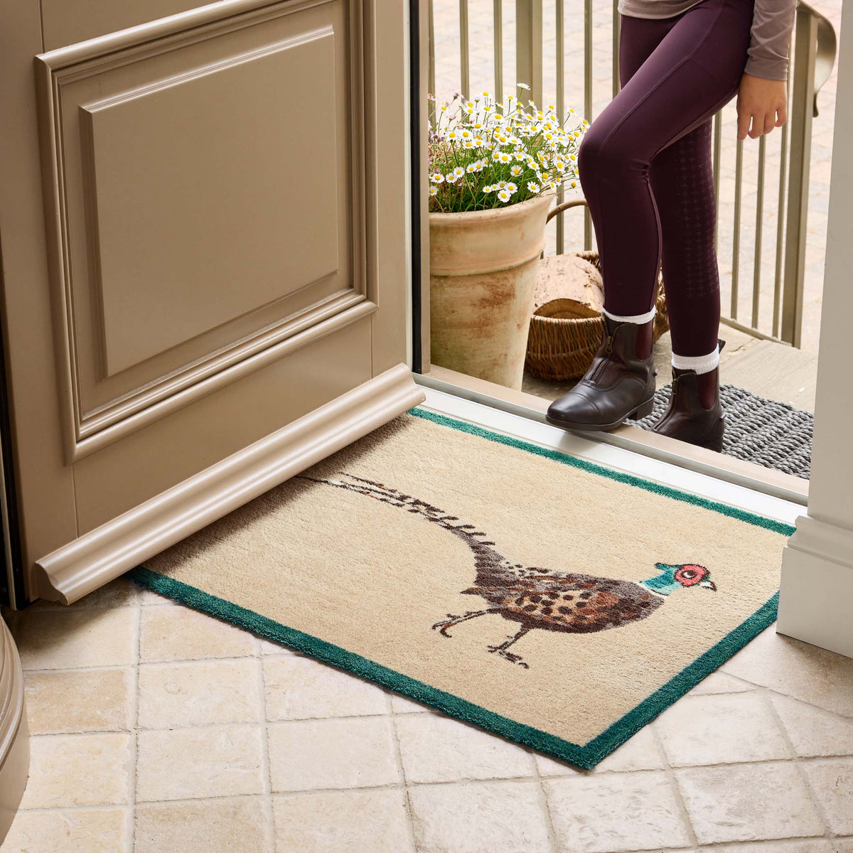 A rectangular entryway doormat with a pheasant illustration and teal border, shown on a tiled floor at a doorway with a person standing outside.