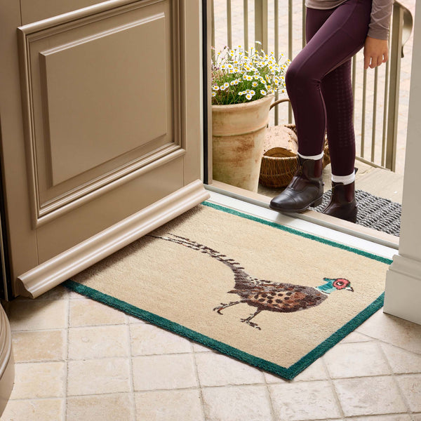 A rectangular entryway doormat with a pheasant illustration and teal border, shown on a tiled floor at a doorway with a person standing outside.