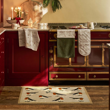 A kitchen scene with a beige doormat featuring a robin pattern in the foreground, with matching patterned towels and oven mitts hanging on a red stove and cabinets.