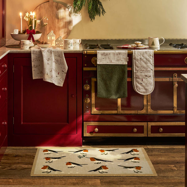 A kitchen scene with a beige doormat featuring a robin pattern in the foreground, with matching patterned towels and oven mitts hanging on a red stove and cabinets.