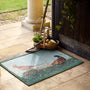 A rectangular doormat with a pheasant hunting scene, placed on a stone patio in front of a wooden door. A basket of produce, a garden tool, and a column are visible in the background.