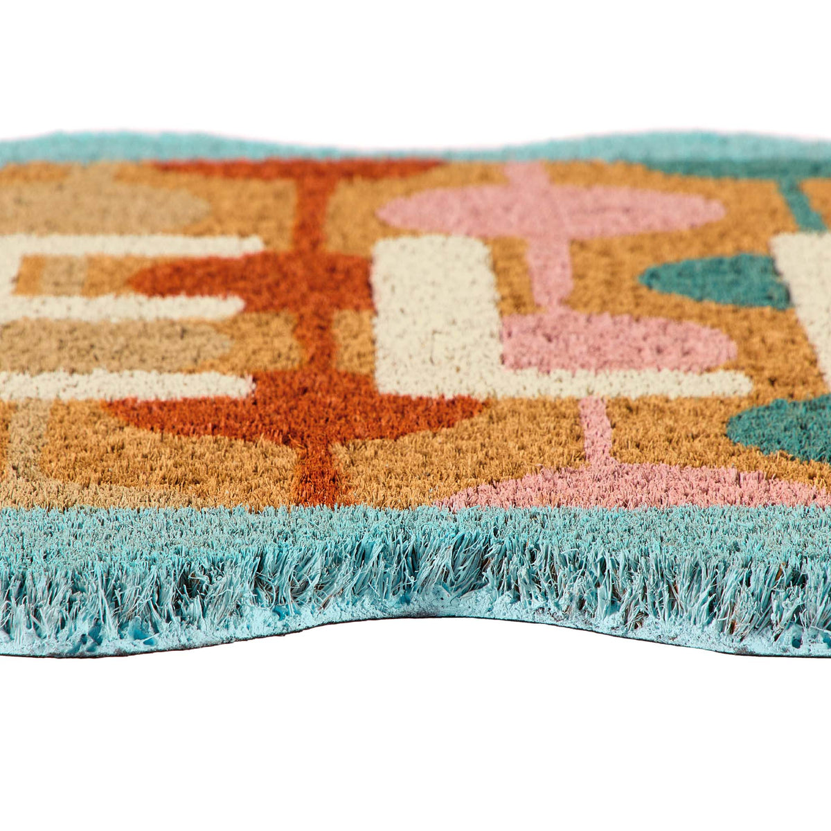 A close-up of a coir doormat with a retro geometric pattern in orange, beige, pink, and teal, featuring a light blue fringe along the bottom edge.