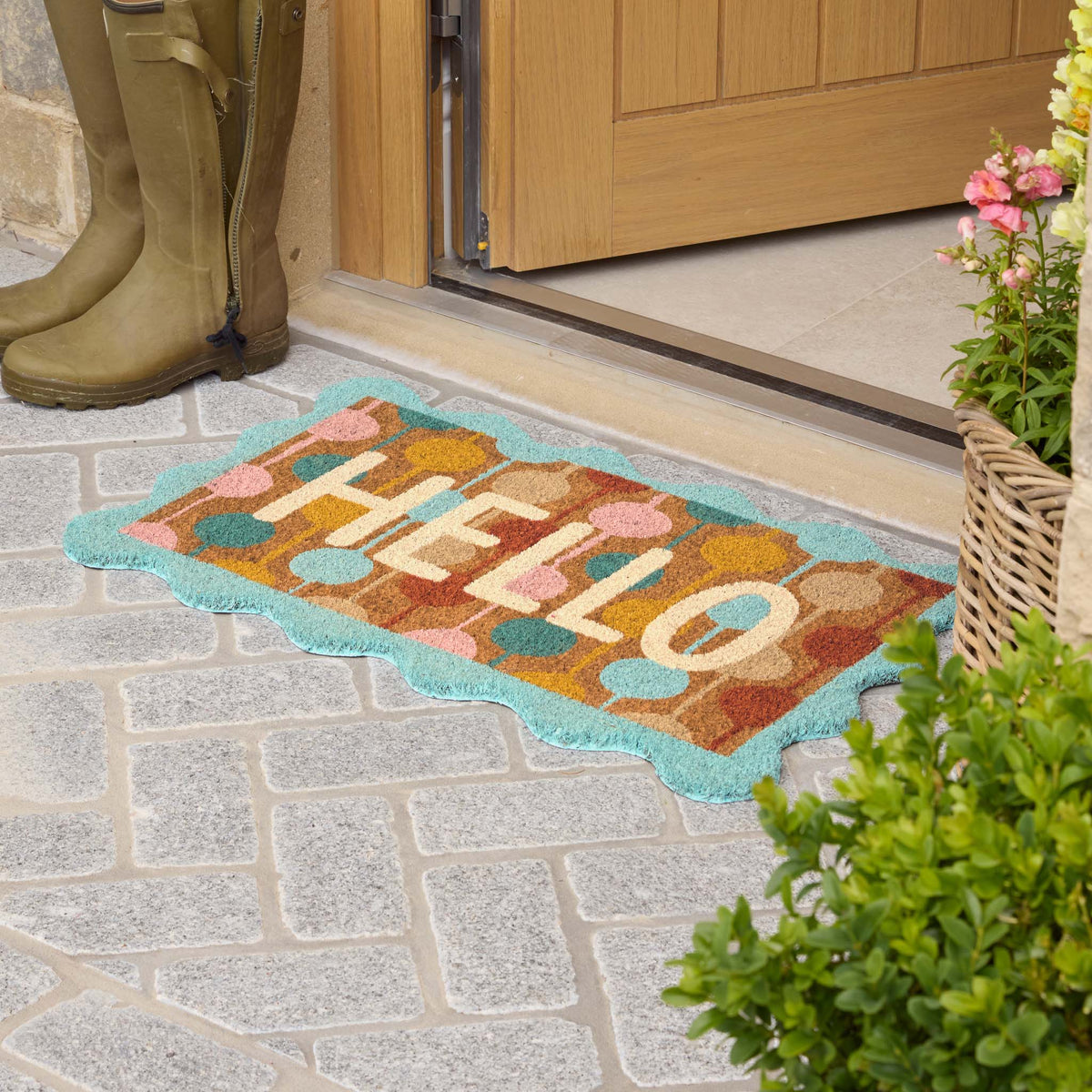 A colorful doormat with the word 'HELLO' in cream letters, surrounded by pastel fruit and a turquoise border, placed on a stone patio in front of a wooden door. Olive green rain boots and potted plants are visible nearby.