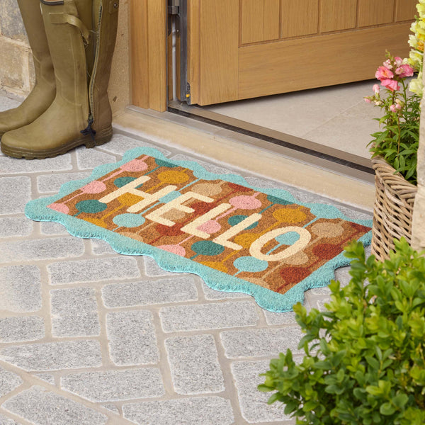 A colorful doormat with the word 'HELLO' in cream letters, surrounded by pastel fruit and a turquoise border, placed on a stone patio in front of a wooden door. Olive green rain boots and potted plants are visible nearby.