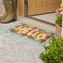 A colorful doormat with the word 'HELLO' in cream letters, surrounded by pastel fruit and a turquoise border, placed on a stone patio in front of a wooden door. Olive green rain boots and potted plants are visible nearby.