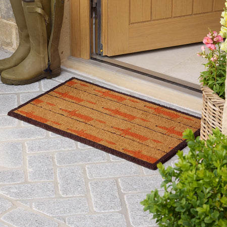 A rectangular coir doormat with a rustic orange and brown geometric pattern, placed on a gray stone patio in front of a wooden door. A pair of olive green rain boots is visible on the left, and a potted plant with pink flowers is on the right.