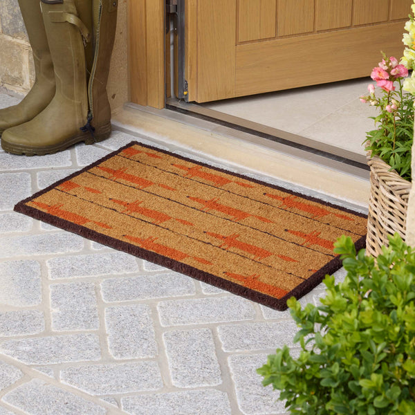 A rectangular coir doormat with a rustic orange and brown geometric pattern, placed on a gray stone patio in front of a wooden door. A pair of olive green rain boots is visible on the left, and a potted plant with pink flowers is on the right.