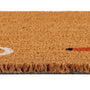 Close-up of a natural coir doormat showing its textured tan-brown fiber pile and fringed edge, with a small portion of a white painted line and a red accent visible on the right.