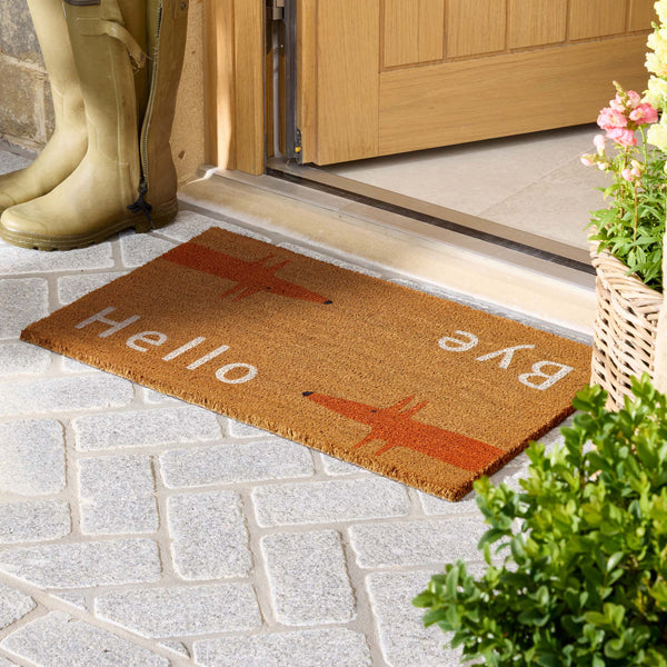 A natural coir doormat with 'Hello' and 'Bye' in white text and orange fox illustrations, placed at a door threshold on a gray stone paver. A person's boots are visible on the left, and a potted plant is on the right.