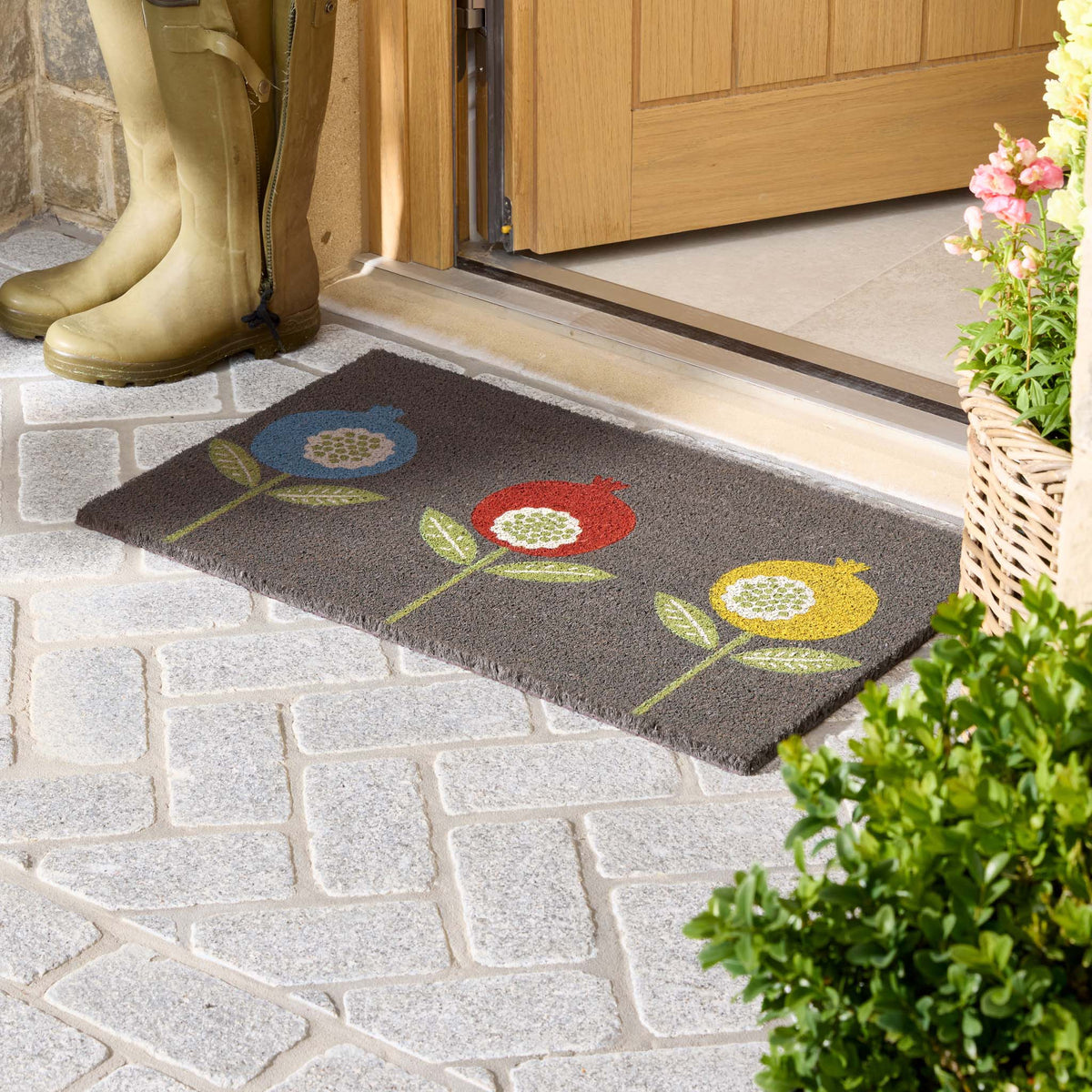 A rectangular dark gray doormat with a colorful flower pattern, placed at a wooden doorway on a stone patio. A pair of tall yellow rain boots stands on the left, and potted plants are visible on the right.