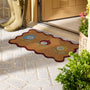 A decorative doormat with a scalloped red border and floral motifs in blue, red, and green, placed on a stone entryway floor near a wooden door, with a person's boots visible at the top left.