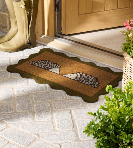 A rectangular doormat with a scalloped dark border and a central hedgehog design, placed on a stone entryway floor near a wooden door. A pair of tall beige rain boots stands nearby, with potted greenery and flowers at the right edge.