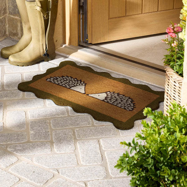 A rectangular doormat with a scalloped dark border and a central hedgehog design, placed on a stone entryway floor near a wooden door. A pair of tall beige rain boots stands nearby, with potted greenery and flowers at the right edge.