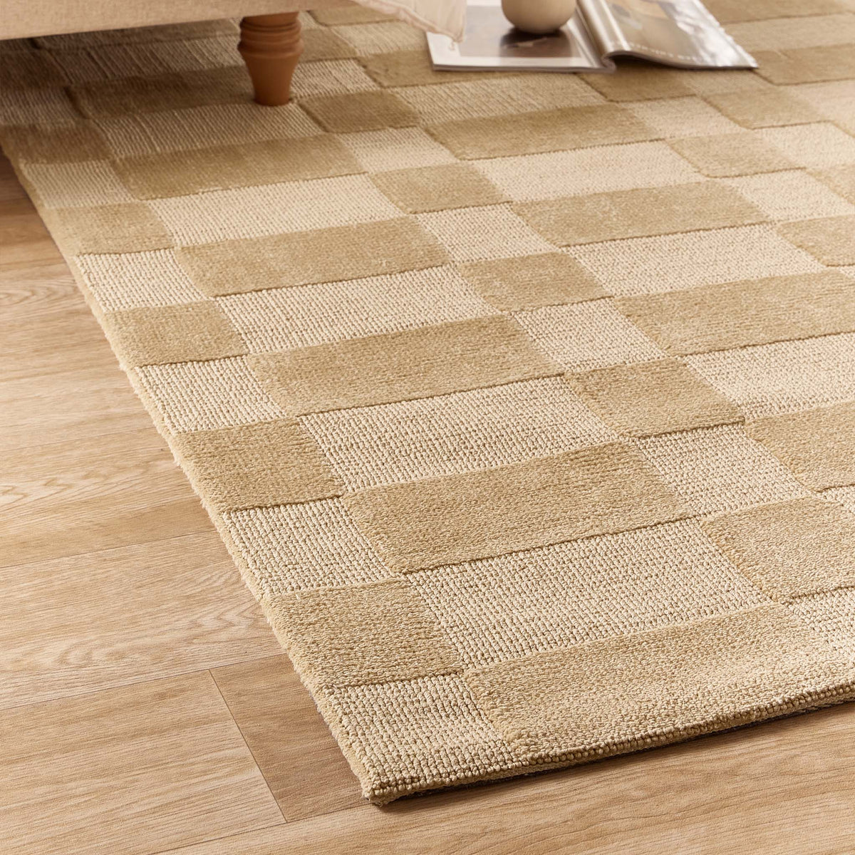 A woven area rug with a beige and tan checkerboard pattern, shown on a light wood floor in a room setting with a table leg and magazine visible.