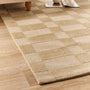 A woven area rug with a beige and tan checkerboard pattern, shown on a light wood floor in a room setting with a table leg and magazine visible.