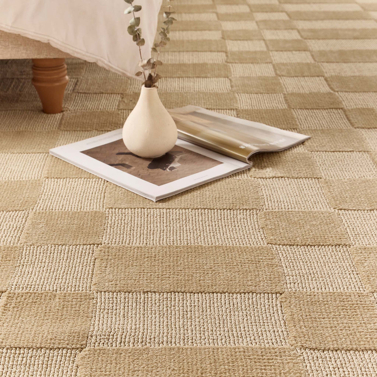 A close-up of a woven area rug with a checkerboard pattern in natural beige and light tan tones, styled with a small white vase, books, and a bed leg in the background.