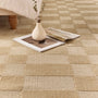 A close-up of a woven area rug with a checkerboard pattern in natural beige and light tan tones, styled with a small white vase, books, and a bed leg in the background.