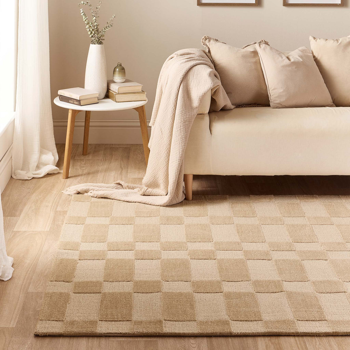 A beige and tan checkered area rug styled in a modern living room setting with a cream sofa, a small side table, and a draped throw blanket.