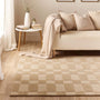 A beige and tan checkered area rug styled in a modern living room setting with a cream sofa, a small side table, and a draped throw blanket.
