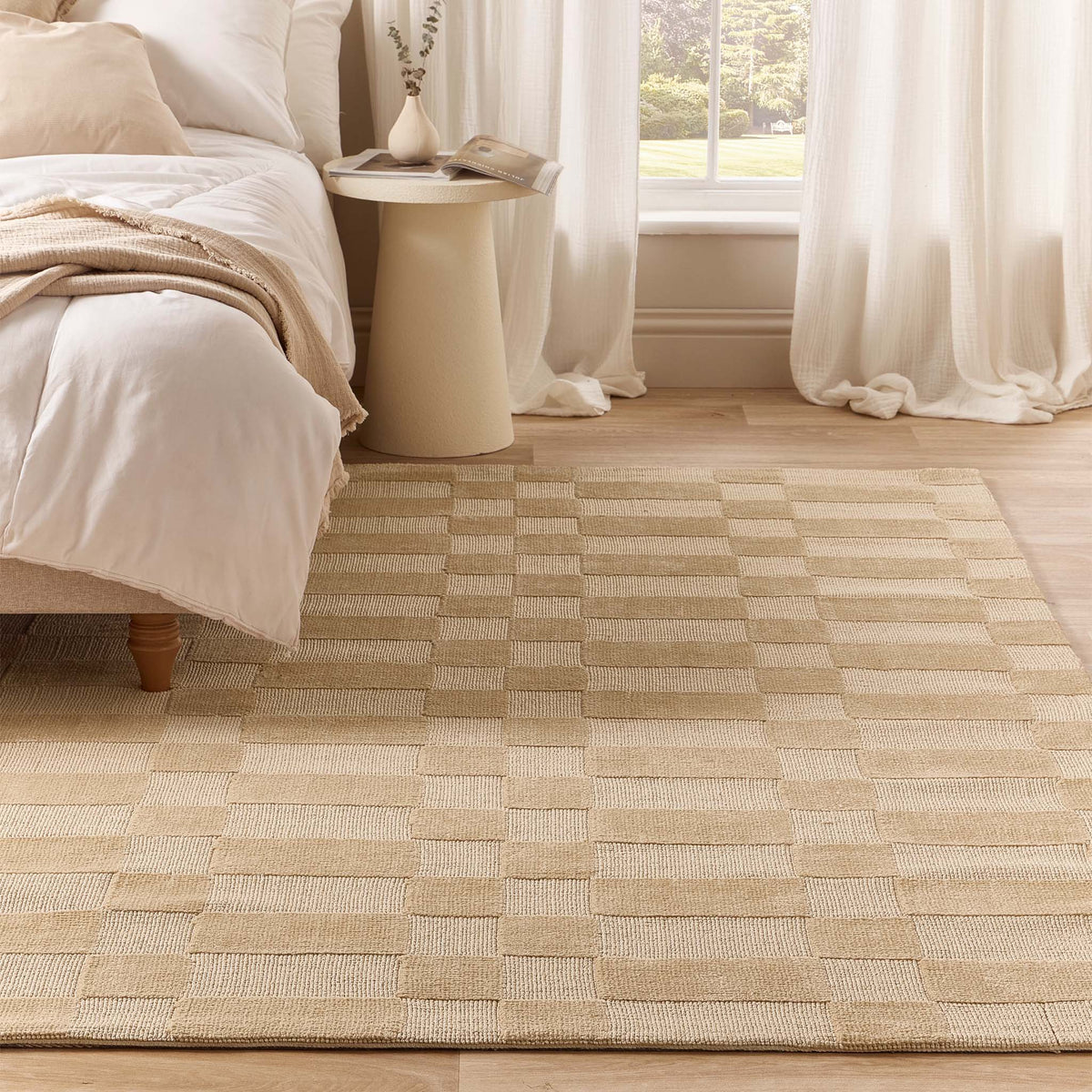 A beige and cream checkered woven area rug styled in a bright, modern bedroom next to a bed and a small side table.