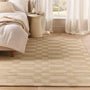 A beige and cream checkered woven area rug styled in a bright, modern bedroom next to a bed and a small side table.