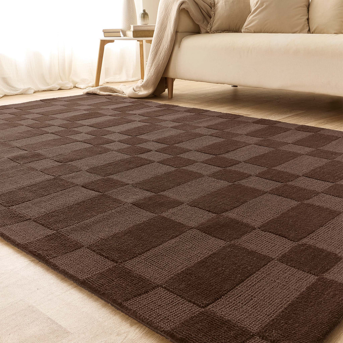 A rectangular area rug with a dark brown and medium brown checkerboard pattern, shown in a brightly lit living room setting with a sofa and side table in the background.