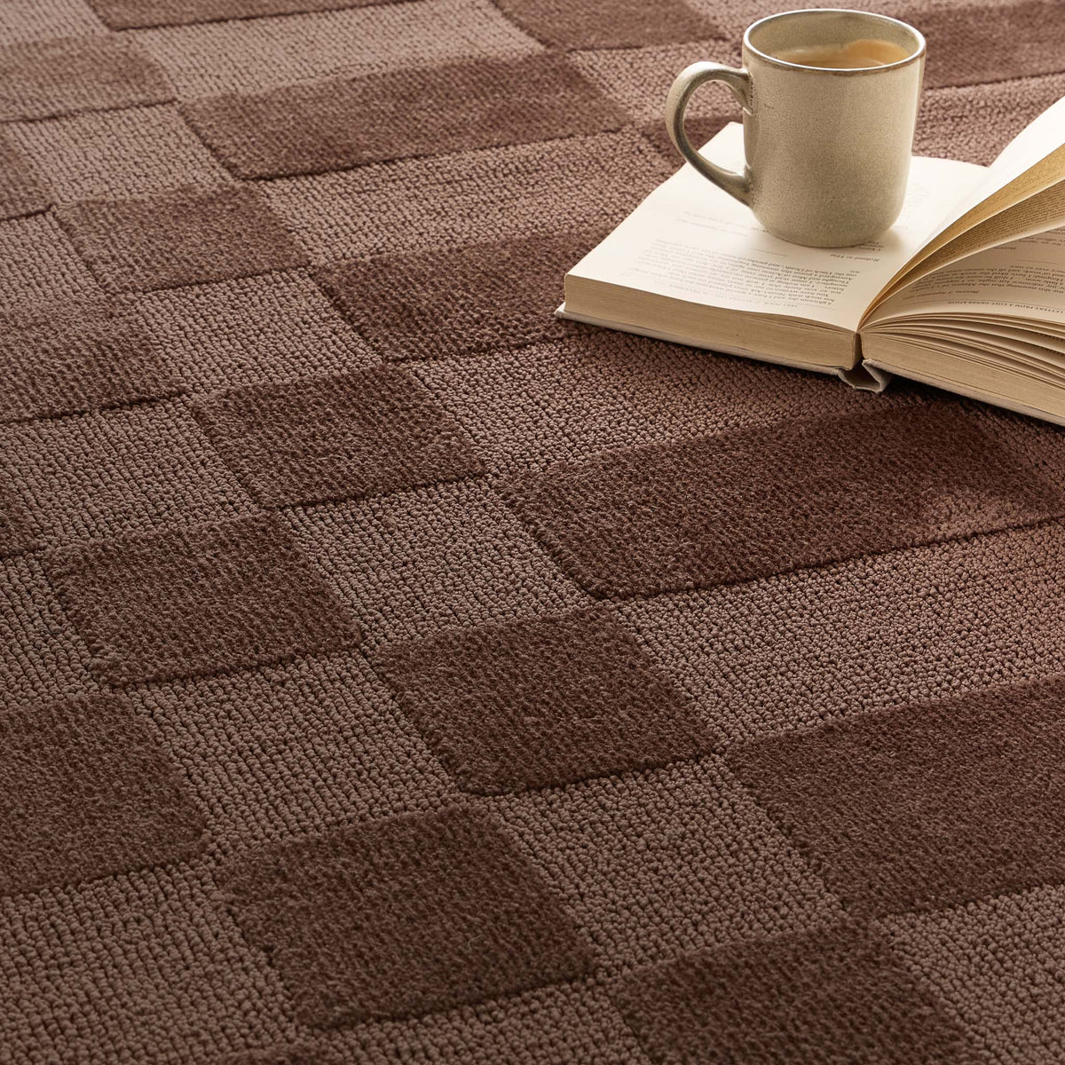 A close-up of a textured brown carpet with a raised checkerboard pattern, styled with an open book and a ceramic mug in the upper right corner.