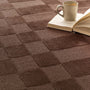 A close-up of a textured brown carpet with a raised checkerboard pattern, styled with an open book and a ceramic mug in the upper right corner.