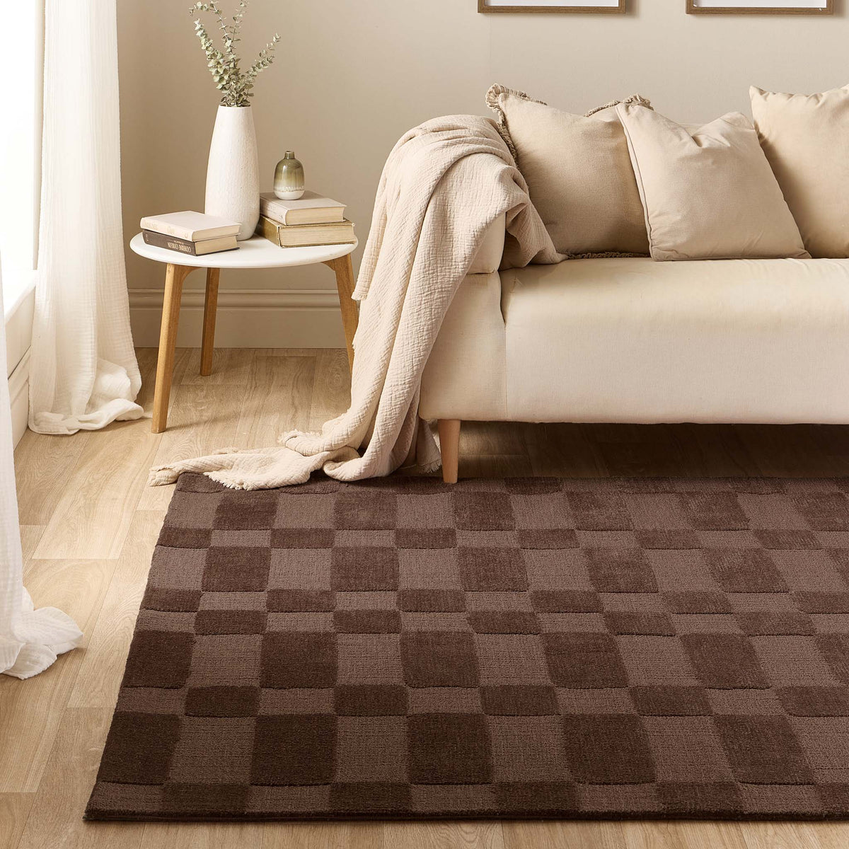 A brown checkered area rug laid on a light wood floor in a styled living room with a cream sofa, pillows, a side table, and a draped throw blanket.