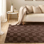 A brown checkered area rug laid on a light wood floor in a styled living room with a cream sofa, pillows, a side table, and a draped throw blanket.
