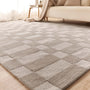 A rectangular area rug with a woven checkerboard pattern in neutral beige and taupe tones, shown on a light wood floor in a bright living room with a sofa and side table visible.