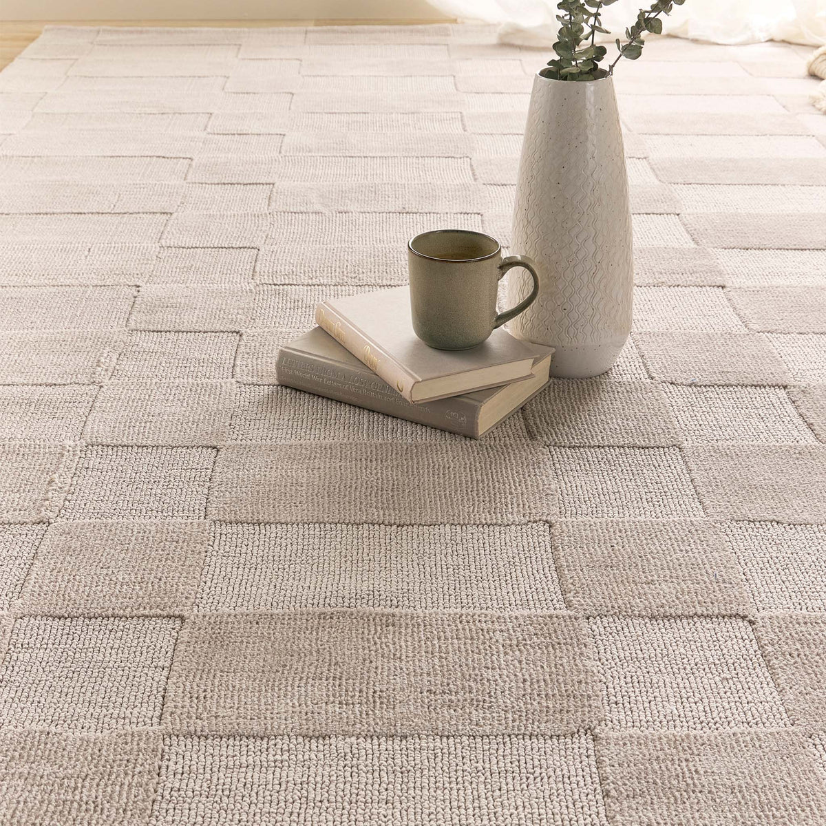 A light beige woven area rug with a textured block pattern, styled in a cozy room setting with a stack of books, a green ceramic mug, and a white textured vase on top.